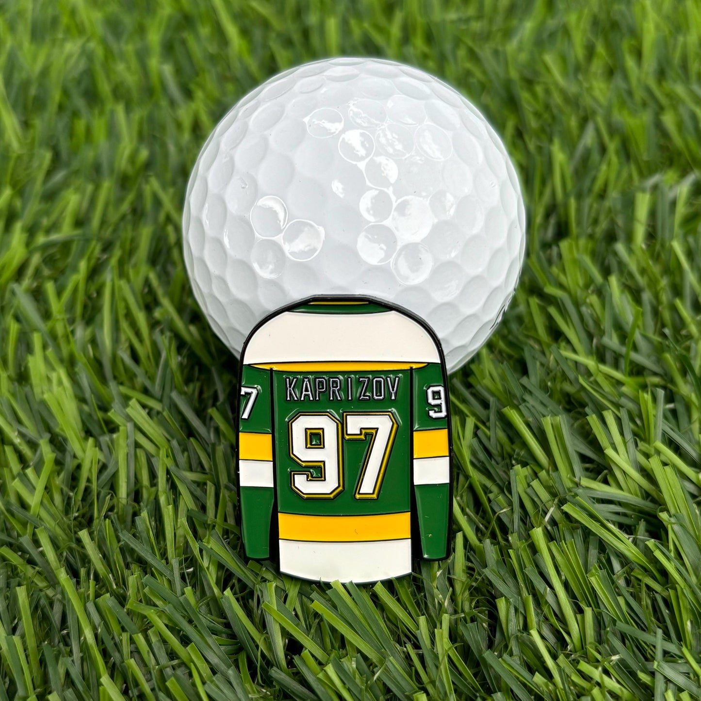 Kirill Kaprizov golf ball marker featuring jersey design, resting on grass with golf ball in background.